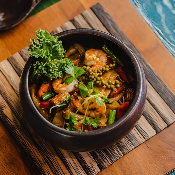 Thai drunken noodles with shrimp, green peppercorns, and vegetables in a dark bowl on a wooden placemat.