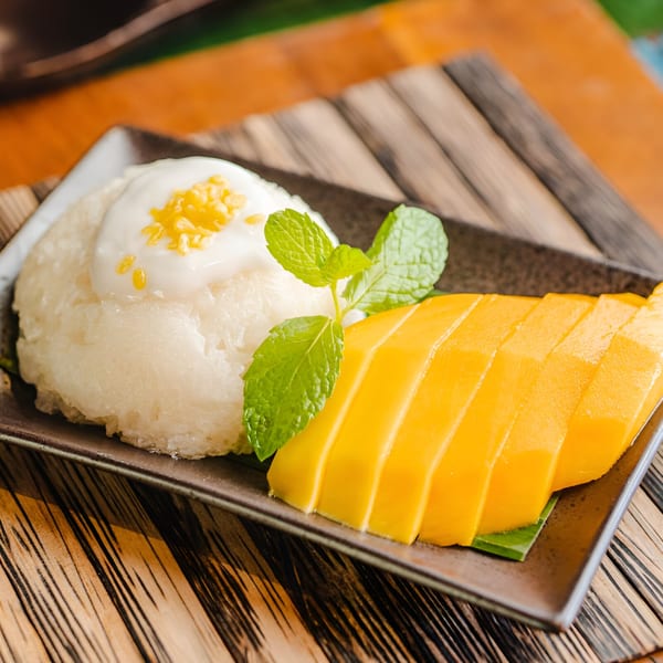 Thai mango sticky rice dessert with coconut cream, sesame seeds, and mint on a dark plate.