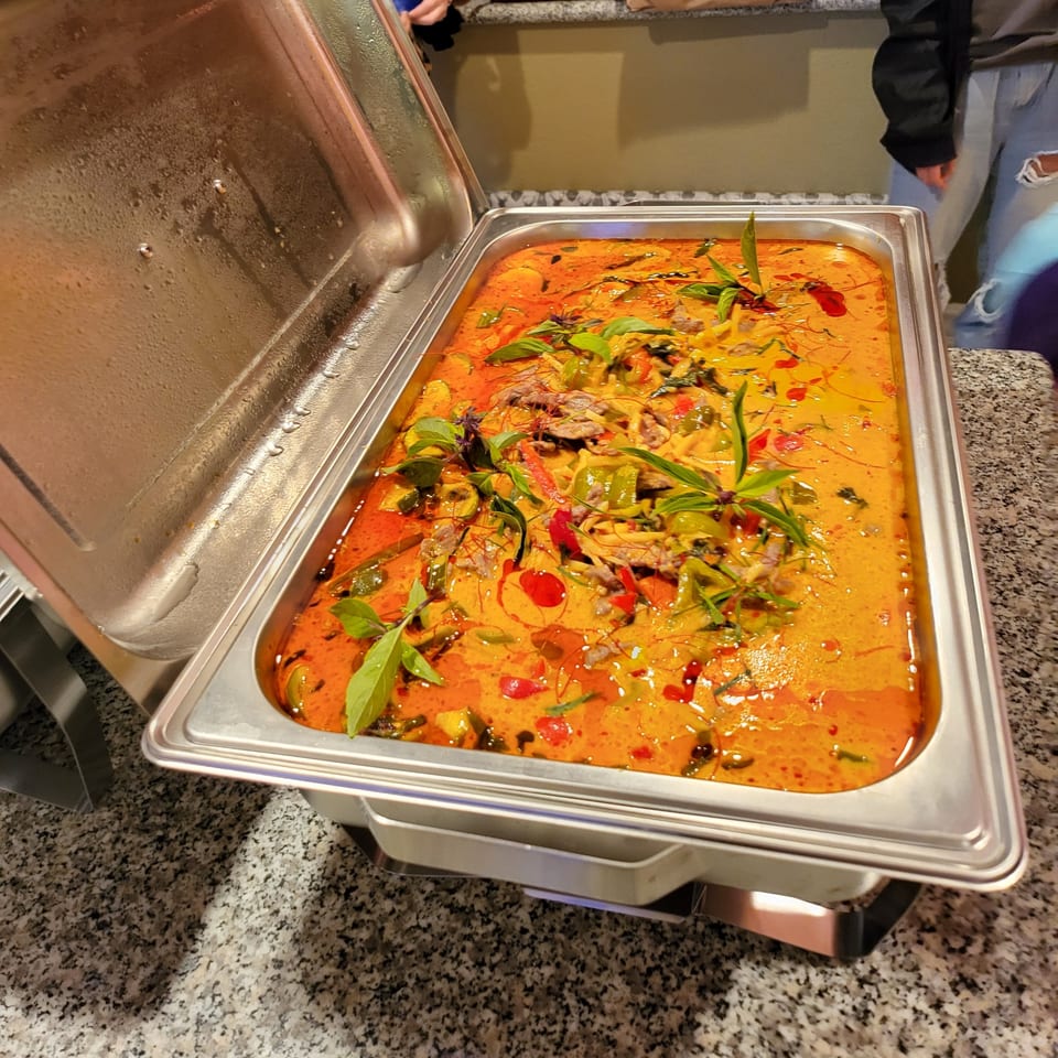 A large buffet tray of vibrant red and orange Thai curry with meat, vegetables, and basil.