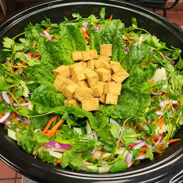 A vibrant Thai salad with crispy tofu, fresh lettuce, cilantro, red onion, carrots, and cucumber in a black bowl.
