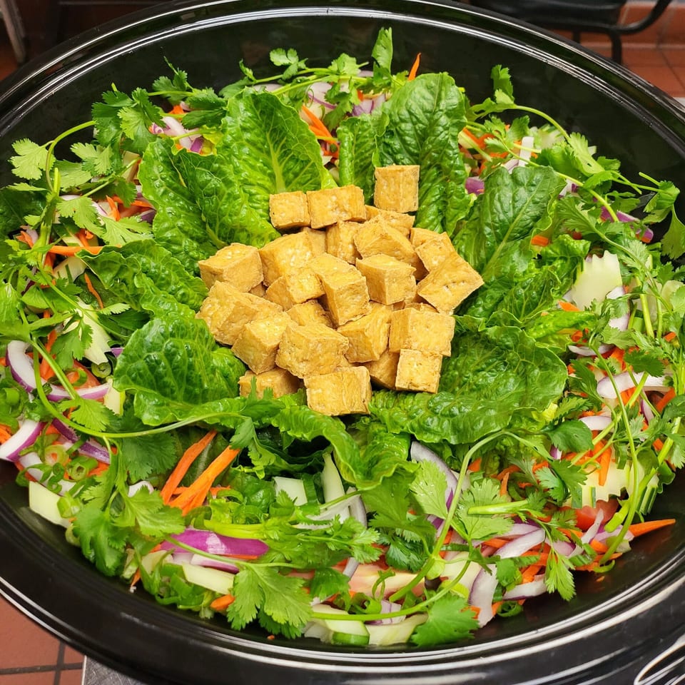 A vibrant Thai salad with crispy tofu, fresh lettuce, cilantro, red onion, carrots, and cucumber in a black bowl.