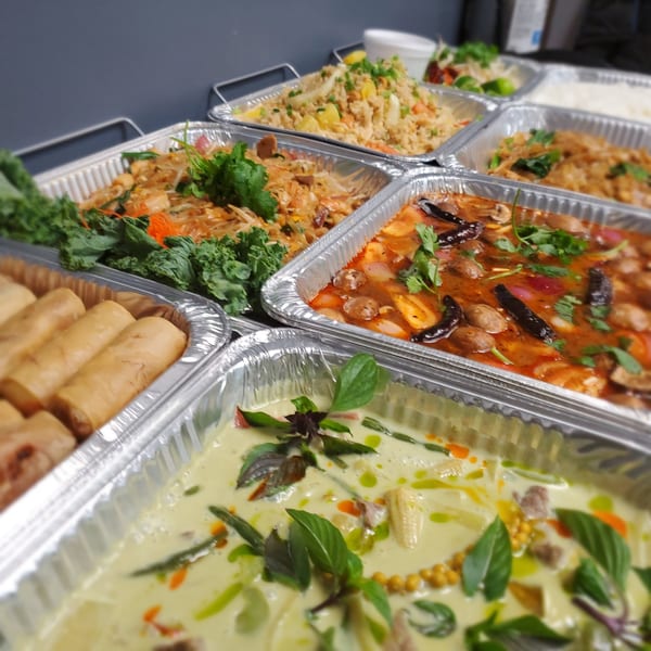 A catering spread of Thai dishes including spring rolls, noodles, curries, and fried rice in foil containers.
