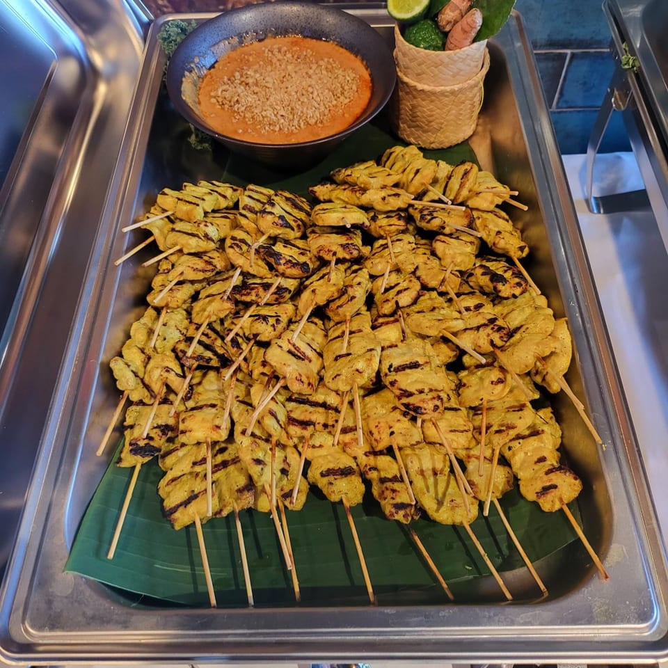 Grilled chicken satay skewers with peanut sauce and garnishes in a metal serving tray.