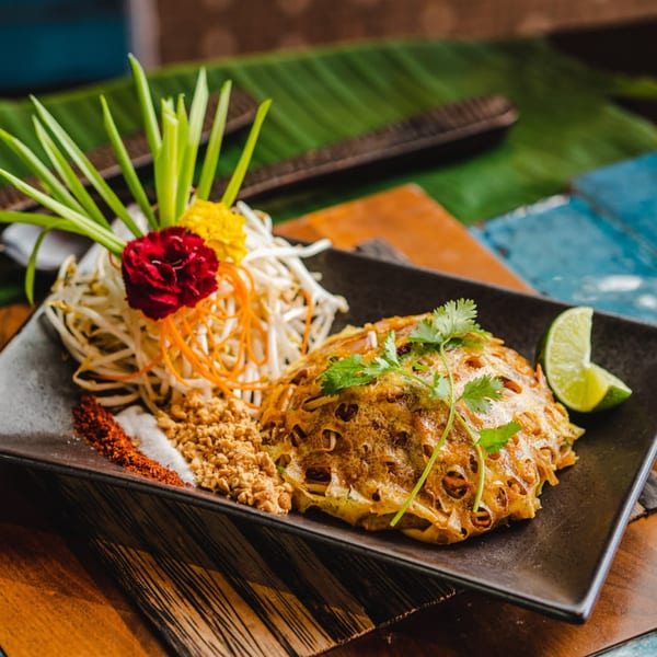 A beautifully plated Pad Thai with a delicate egg net, fresh garnishes, and lime on a dark rectangular dish.