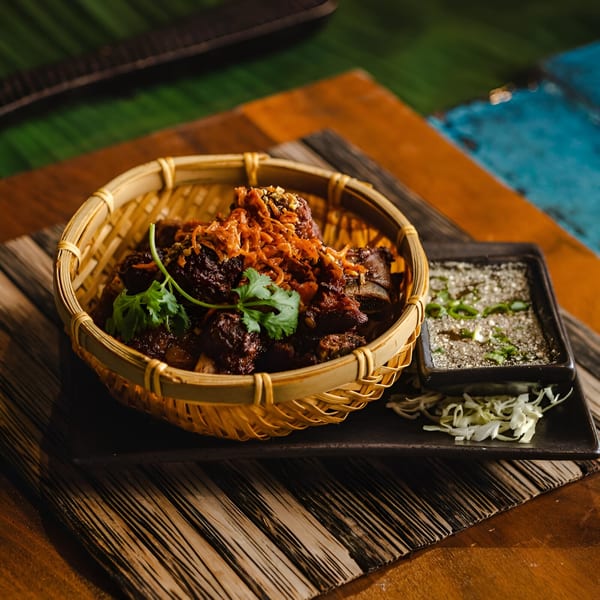 Thai beef ribs in a woven basket with cilantro, fried shallots, and a side of dipping sauce.