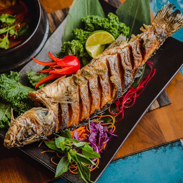 Crispy whole fried fish garnished with lime, red chili, kale, and herbs on a black plate.