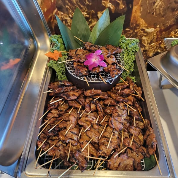 Grilled Thai pork skewers, Moo Ping, arranged in a chafing dish with a purple orchid garnish.